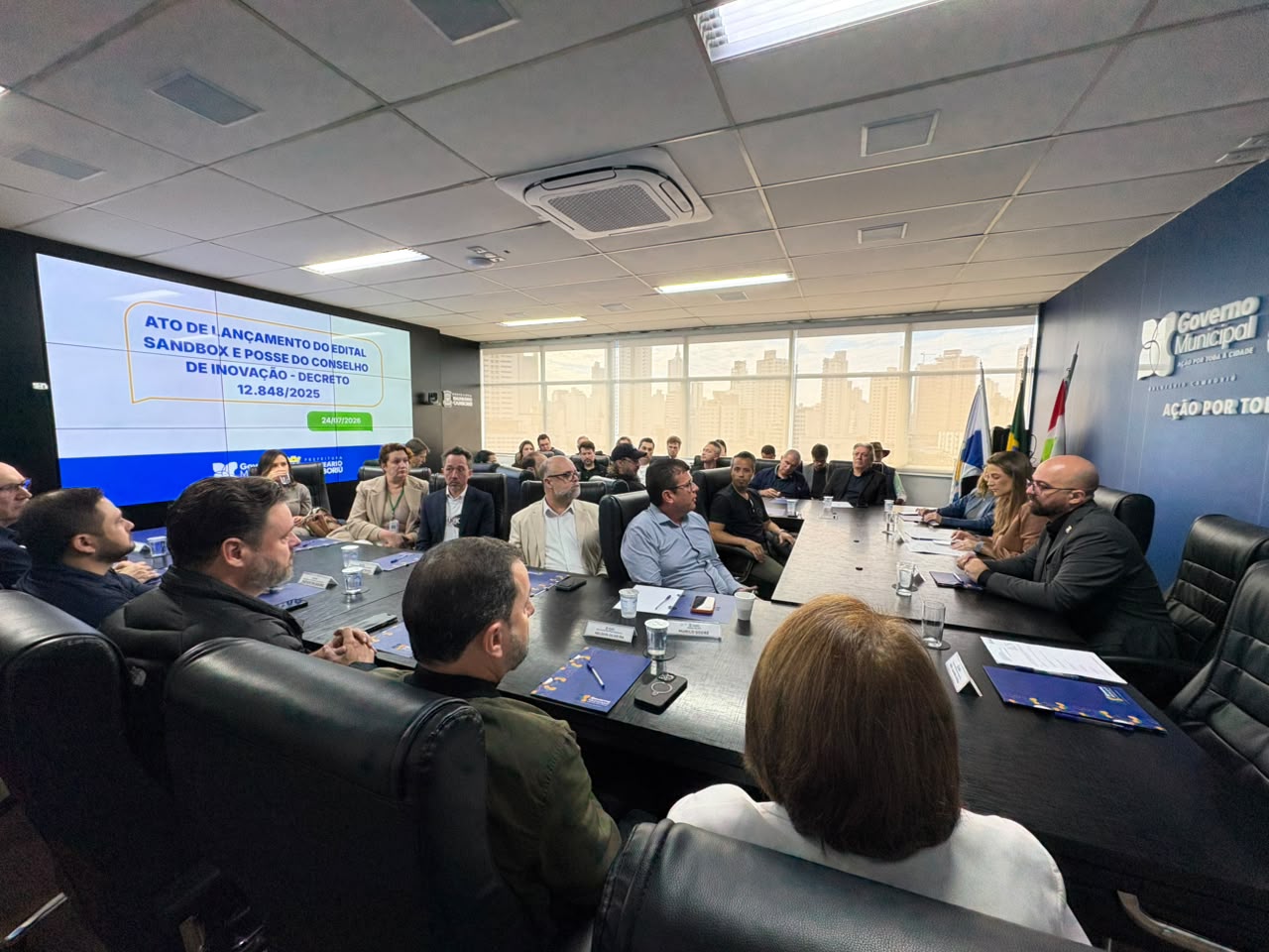 Sessão de posse do Conselho Municipal de Ciência, Tecnologia e Inovação de Balneário Camboriú, com os conselheiros reunidos à mesa e o telão anunciando o ato de lançamento do edital do Sandbox Regulatório.